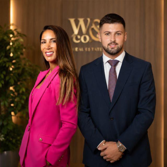 A man in a navy blue suit and a woman in a bright pink blazer standing in front of a warm wood-paneled wall with a gold 'W&Co Real Estate' logo.